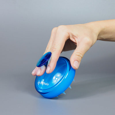 Eine Hand hält eine Dr. Balwi Massagebürste von Dr. Balwi Shop mit kurzen, weichen Borsten zur Kopfhautmassage und zur Unterstützung des Haarwachstums vor einem schlichten grauen Hintergrund.