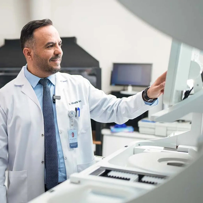 Ein Arzt in weißem Laborkittel und blauer Krawatte lächelt, während er in einem modernen medizinischen Labor Laborgeräte bedient. Im Hintergrund sind Computermonitore und Laborgeräte zu sehen.