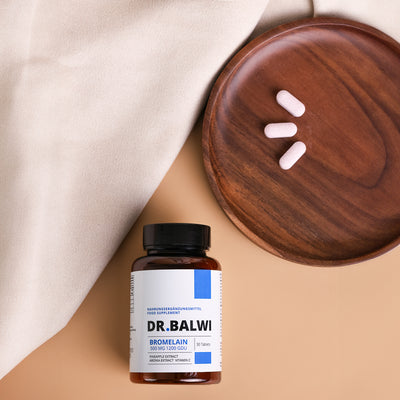 Ein Fläschchen Dr. Balwi Shop Bromelain ruht auf beigem Stoff neben einem Holzteller mit drei rosa Tabletten, die die Vorteile von Ananas Extrakt für alle, die von Haarverlust betroffen sind, aufzeigen.