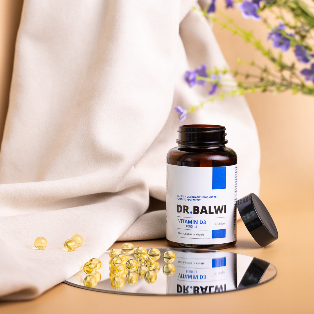 Eine Flasche Dr. Balwi Shop Vitamin D 1000 IE mit offenem Deckel steht auf einer spiegelnden Fläche, weiche Gelkapseln liegen verstreut daneben - ideal, um das Immunsystem zu stärken. Im Hintergrund sind lila Blumen und ein weißes Tuch zu sehen.