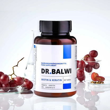 Eine Flasche Biotin & Keratin Tabletten aus dem Dr. Balwi Shop ist neben einem Glasbecher, einem Glas und roten Trauben auf einer weißen Fläche abgebildet.