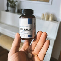 Eine Hand hält eine braune Flasche mit der Aufschrift Biotin & Keratin Tabletten von Dr. Balwi Shop mit einer Tablette auf der Handfläche; ein weißer Tisch und eine unscharfe Wohnungseinrichtung im Hintergrund betonen die Unterstützung für Haarwachstum.