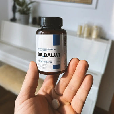 Eine Hand hält eine braune Flasche mit der Aufschrift Biotin & Keratin Tabletten von Dr. Balwi Shop mit einer Tablette auf der Handfläche; ein weißer Tisch und eine unscharfe Wohnungseinrichtung im Hintergrund betonen die Unterstützung für Haarwachstum.