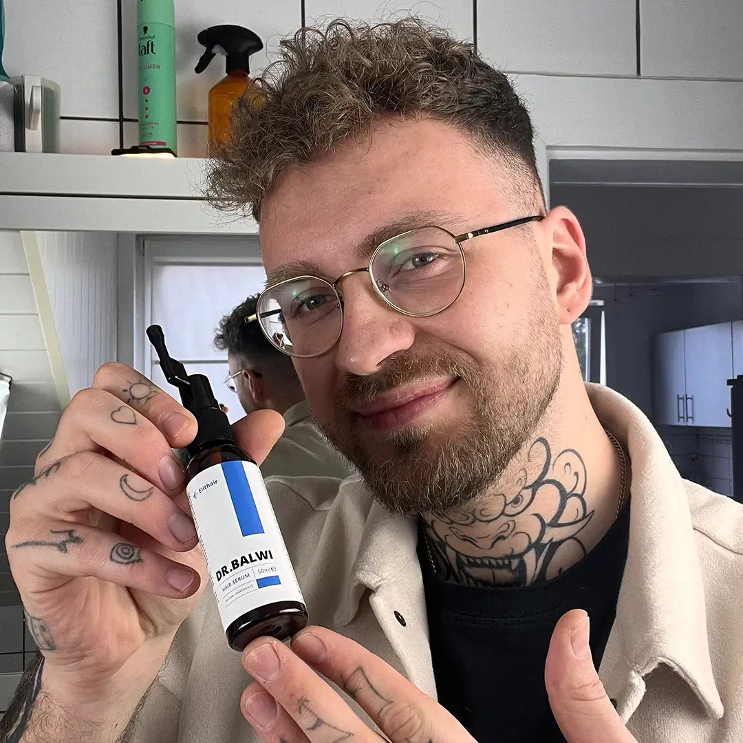 Ein Mann mit Brille und Tattoos lächelt, während er eine Flasche Dr. Balwi Hair Serum aus dem Dr. Balwi Shop in einem modernen Badezimmer mit Regalen und einem Spiegel im Hintergrund hochhält.