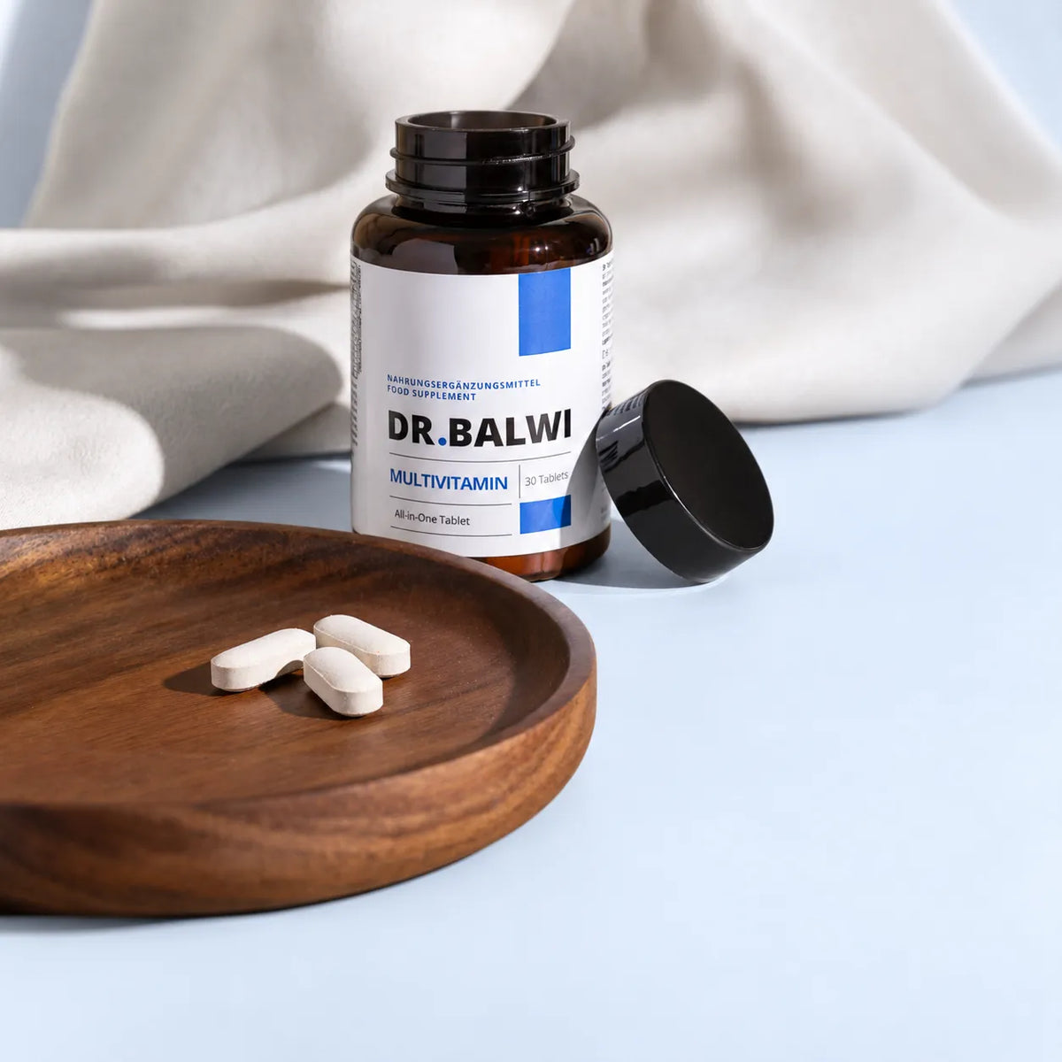 Eine geöffnete Flasche Dr. Balwi Shop Multivitamin Complex mit drei Tabletten, die auf einem kleinen Holzteller liegen.