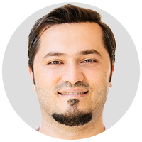 Ein Mann mit kurzen dunklen Haaren und gestutztem Spitzbart lächelt sanft in die Kamera. Er trägt ein helles Hemd und steht vor einem schlichten, hellgrauen Hintergrund.