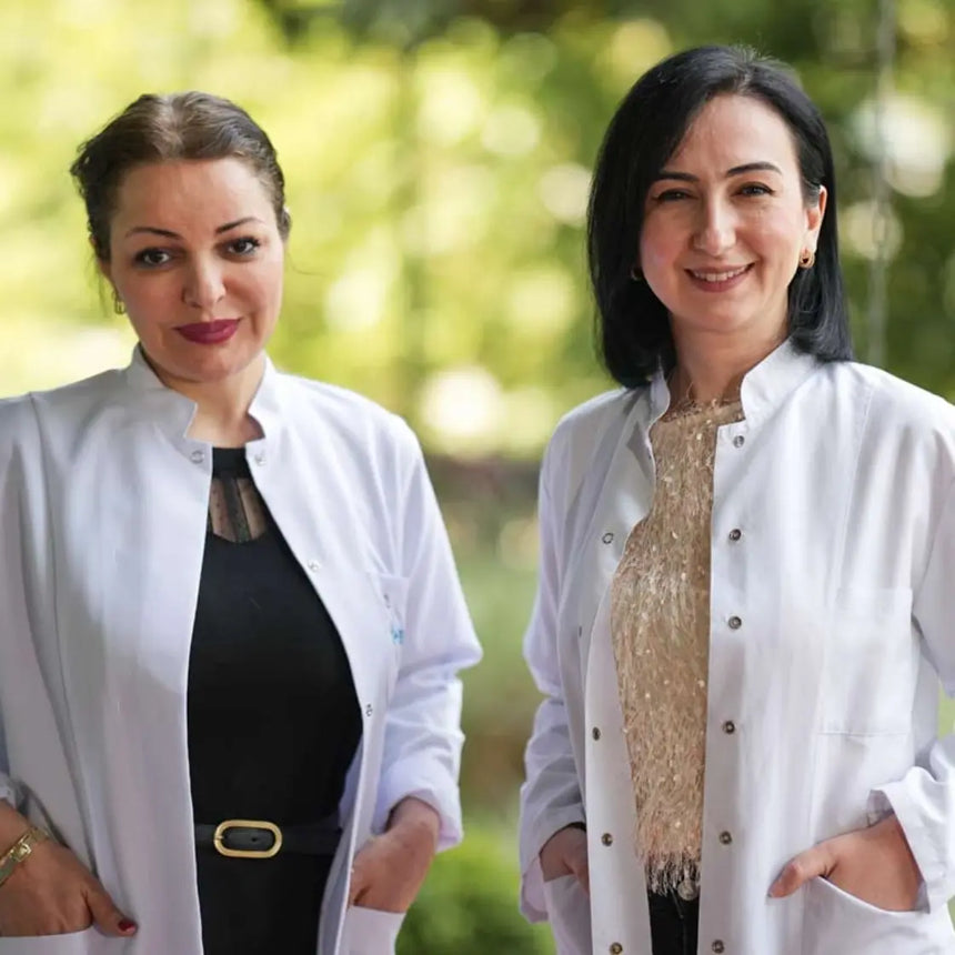 Zwei Frauen in weißen Laborkitteln stehen im Freien und lächeln in die Kamera. Der Hintergrund ist verschwommenes Grün, was an eine Park- oder Gartenkulisse erinnert. Beide haben dunkle Haare und die Hände in den Manteltaschen.